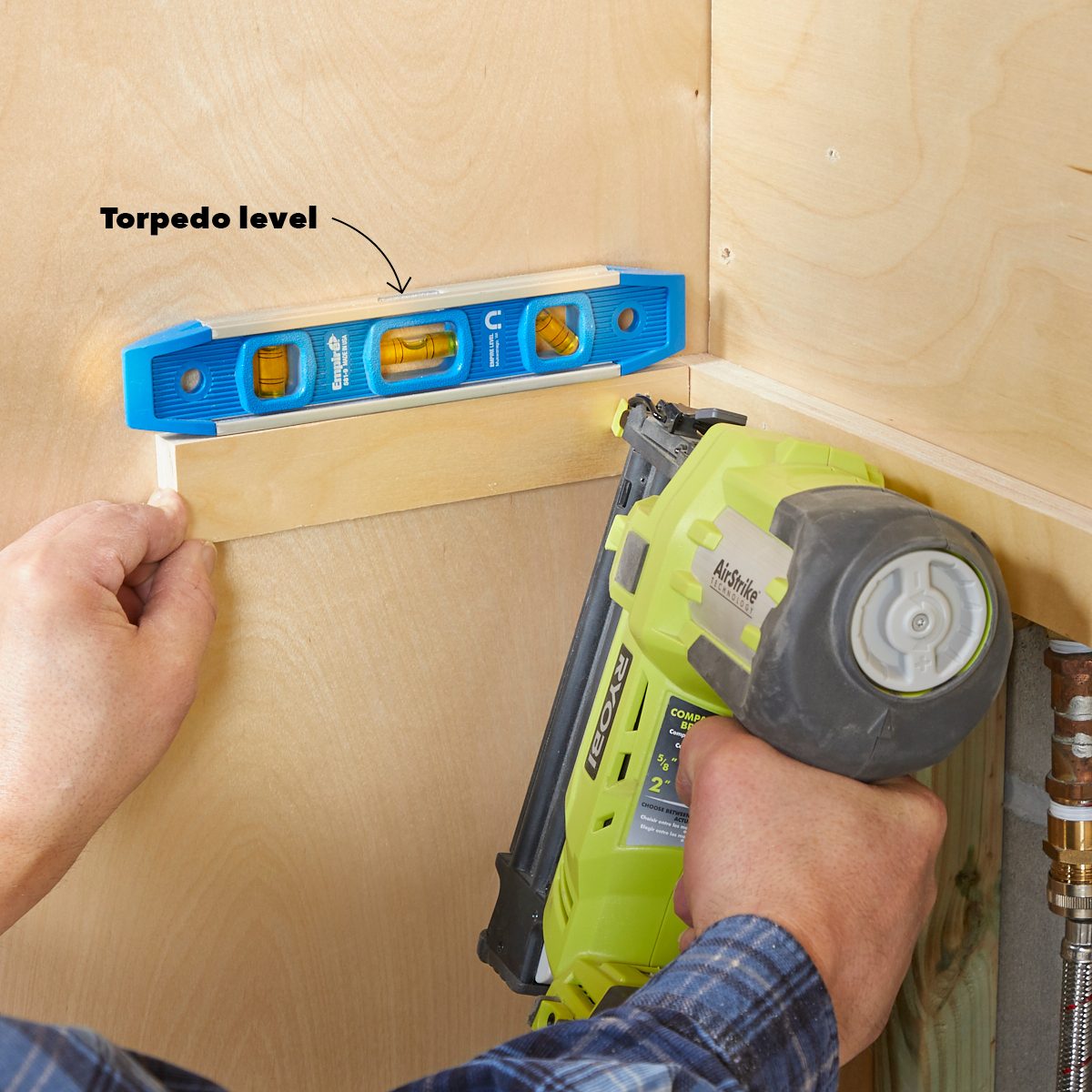 A hand holds a torpedo level against a wooden surface while another hand operates a nail gun, ensuring a precise alignment in a construction setting.