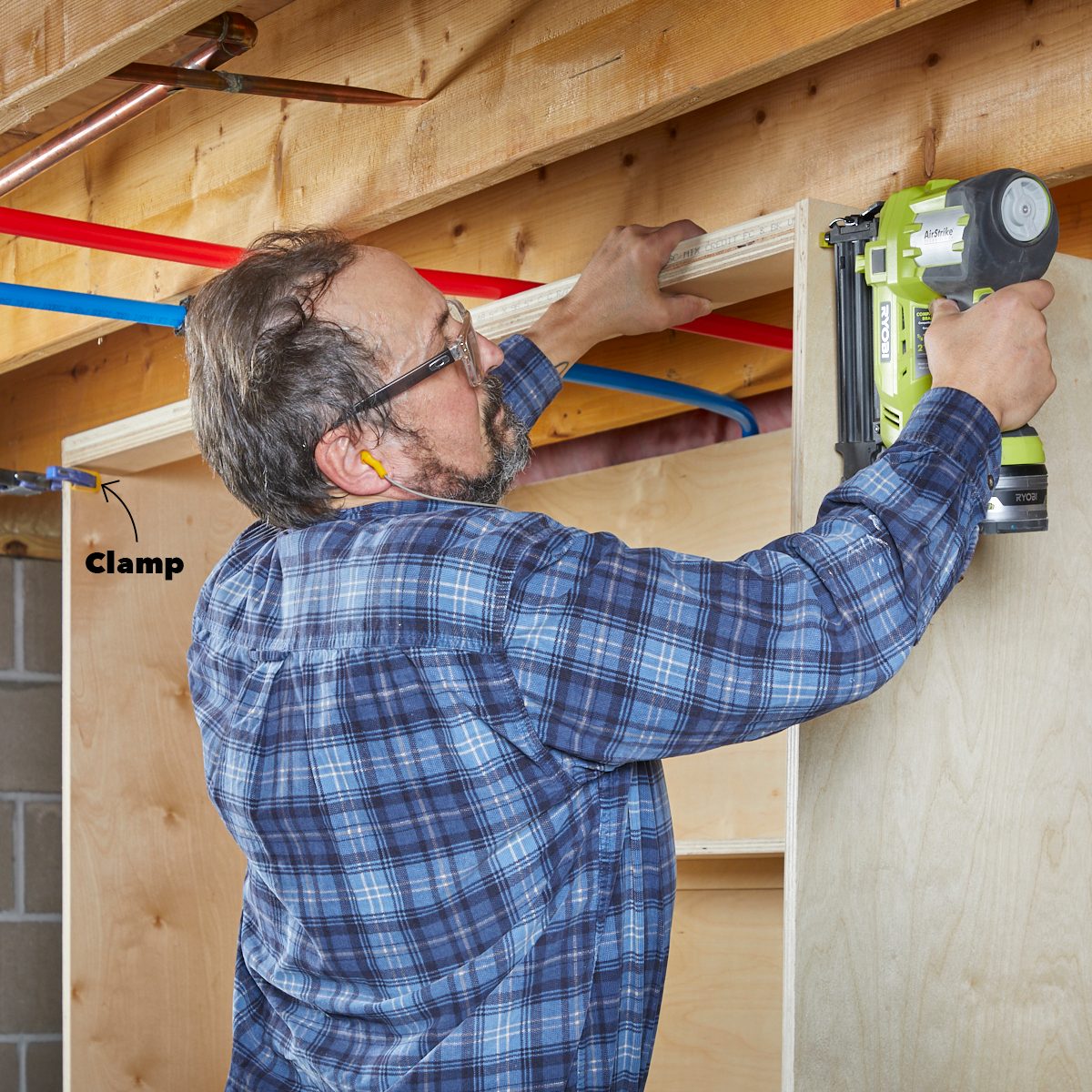 A man uses a nail gun to secure a wooden board to a wall, while a clamp holds it in place under a wooden ceiling framework.