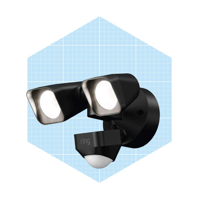 A dual-headed security light is mounted on a wall, illuminating surroundings, while a motion sensor below detects movement. The background features a light blue grid.