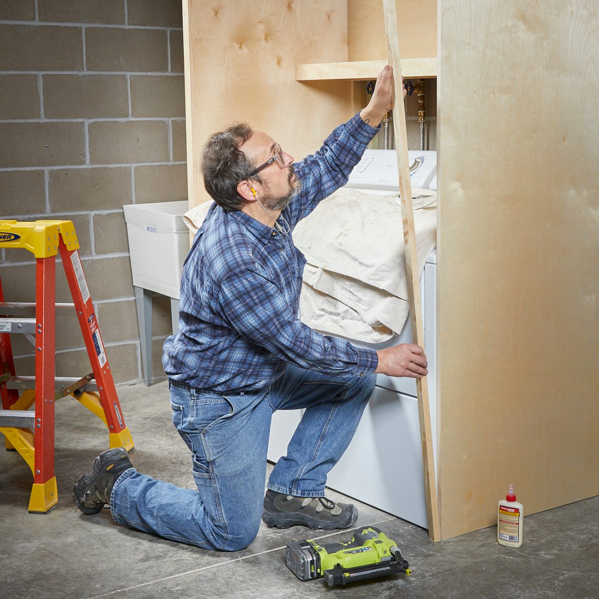 A man kneels, measuring wood against a wall, while working in a garage with a washer and tools nearby, including a ladder and adhesive.