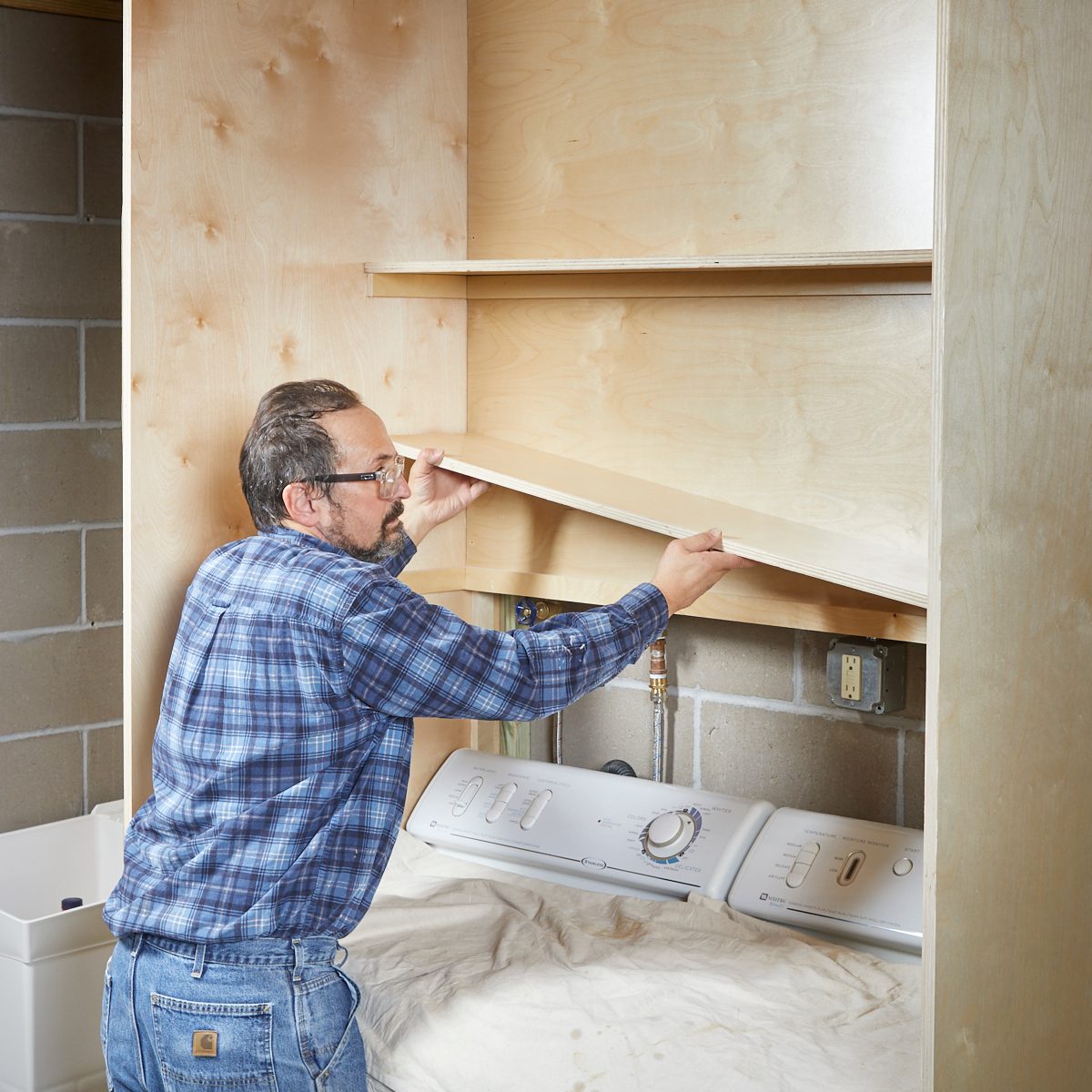 A man installs a shelf in a wooden structure above a laundry appliance in a room with concrete walls and a partially covered floor.
