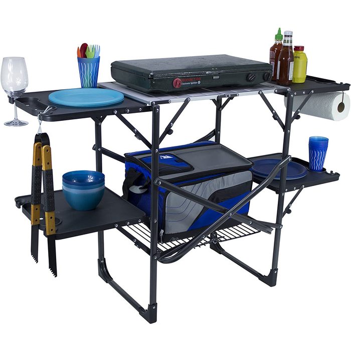 A folding table displays various kitchen items, including plates, bowls, cooking tools, and condiments, set in an organized manner for outdoor cooking or dining.