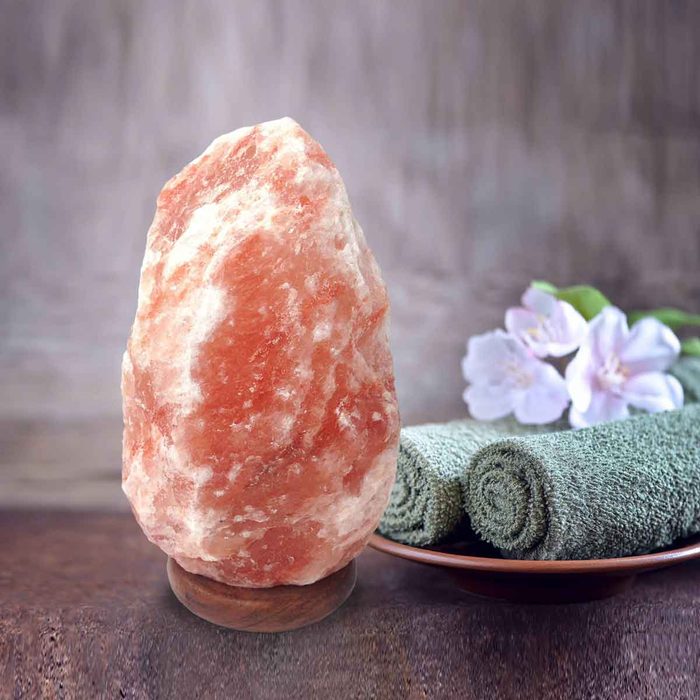A pink salt lamp stands on a wooden base, with rolled green towels and flowers nearby, arranged on a rustic surface.