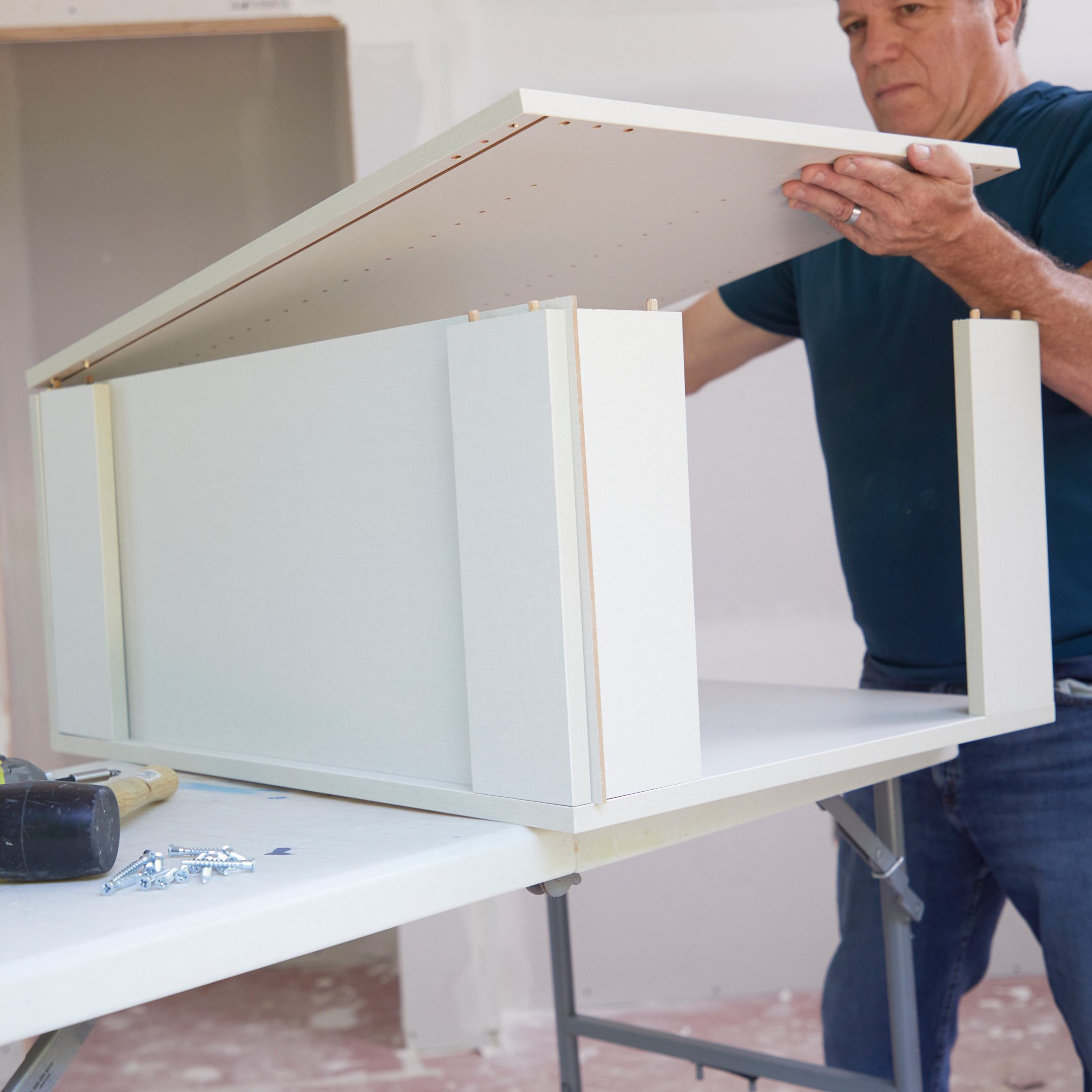 A man assembles furniture by placing the top panel onto a partially constructed cabinet on a table in a room with unfinished walls.