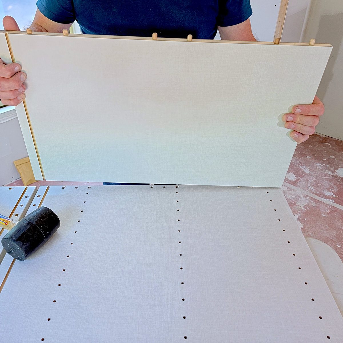 A person holds a large white panel above a work surface with multiple drilled holes, preparing to align it with the holes for assembly.
