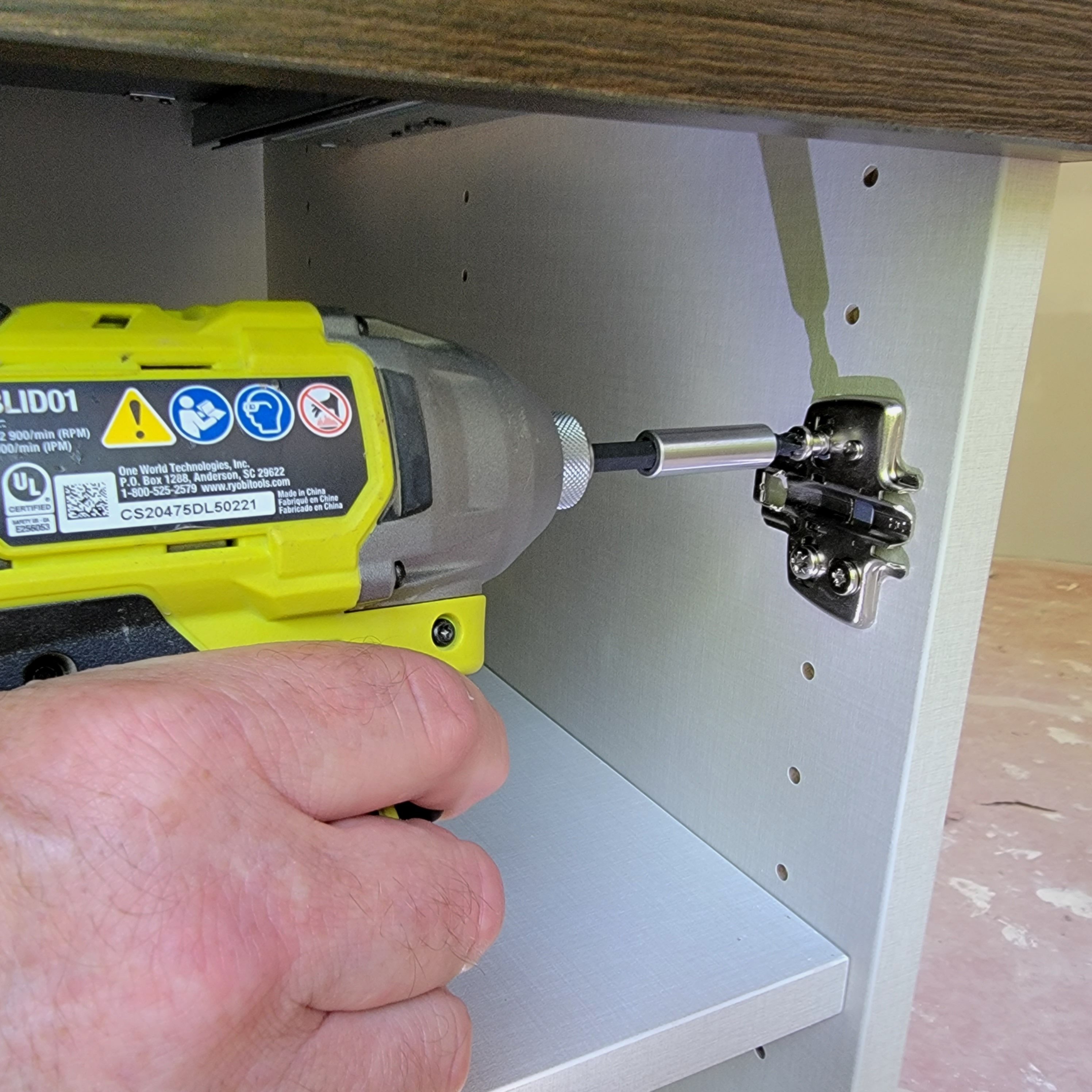 A hand grips a power screwdriver, attaching a metal hinge to the interior panel of a cabinet, surrounded by light-colored materials and a faintly textured surface.