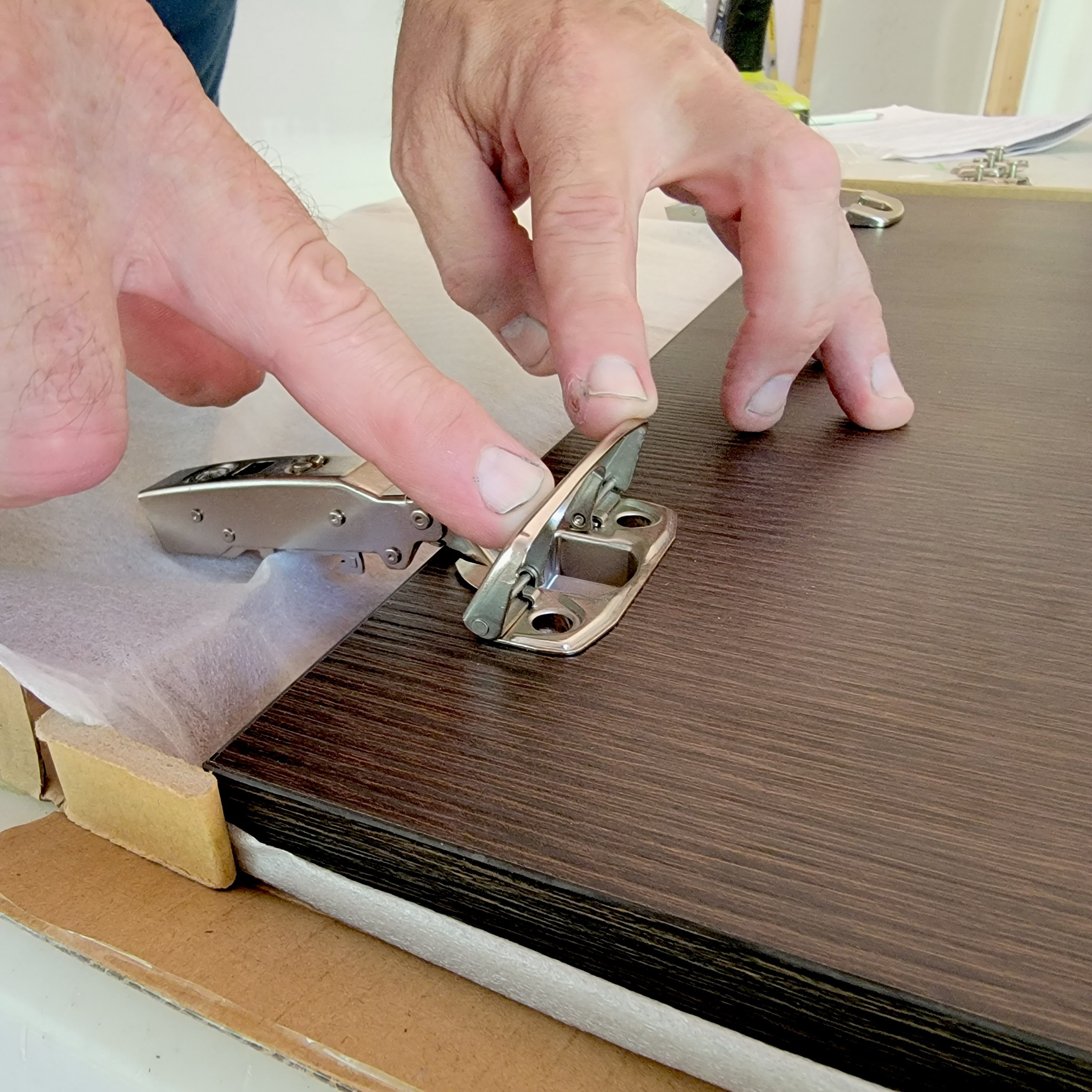 A hand positions a metal hinge on a dark wooden surface while preparing for assembly in a lightly lit interior environment.