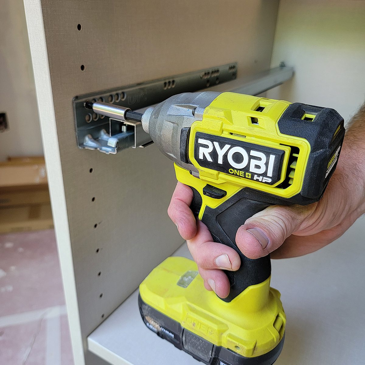 A hand holds a yellow and gray cordless drill, attaching a metal bracket to a wooden surface in a construction environment.
