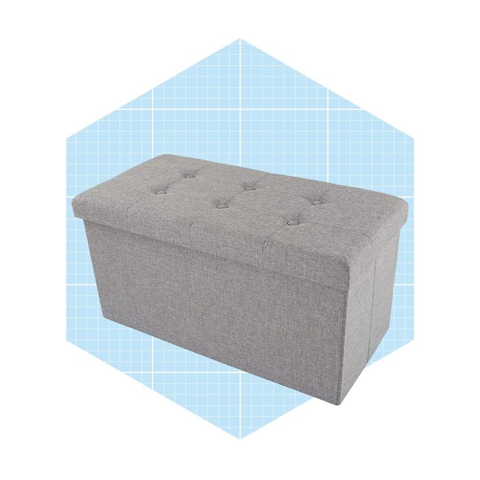 A rectangular gray upholstered ottoman sits on a light blue grid-patterned background, featuring a tufted top and hidden storage inside.