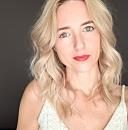A woman with wavy blonde hair and red lips looks directly at the camera, set against a neutral background that enhances her features.