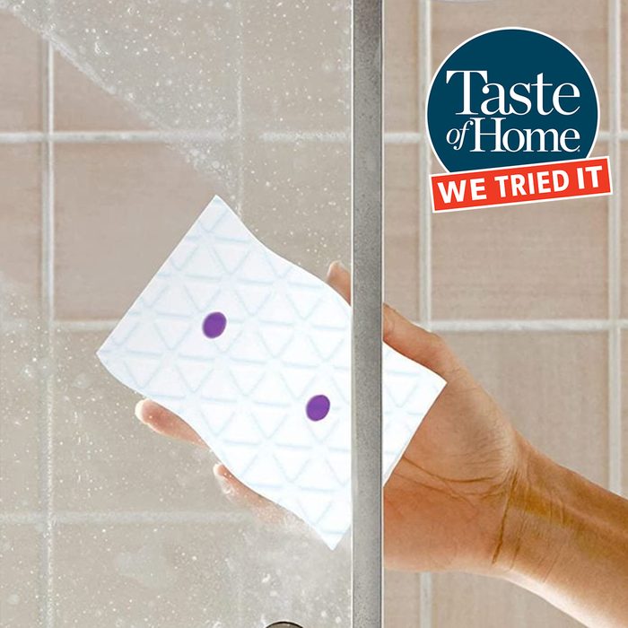 A hand holds a cleaning pad against a steamy shower door, wiping away moisture while surrounded by tiled bathroom walls.