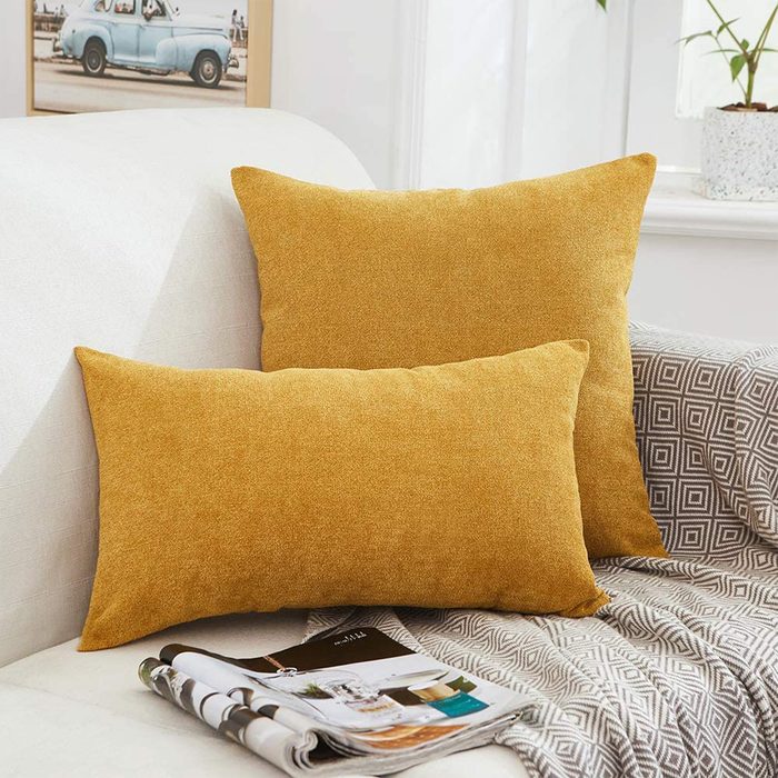 Two golden-yellow pillows rest on a light-colored sofa, accompanied by a folded blanket and a stack of magazines, in a cozy, bright room.