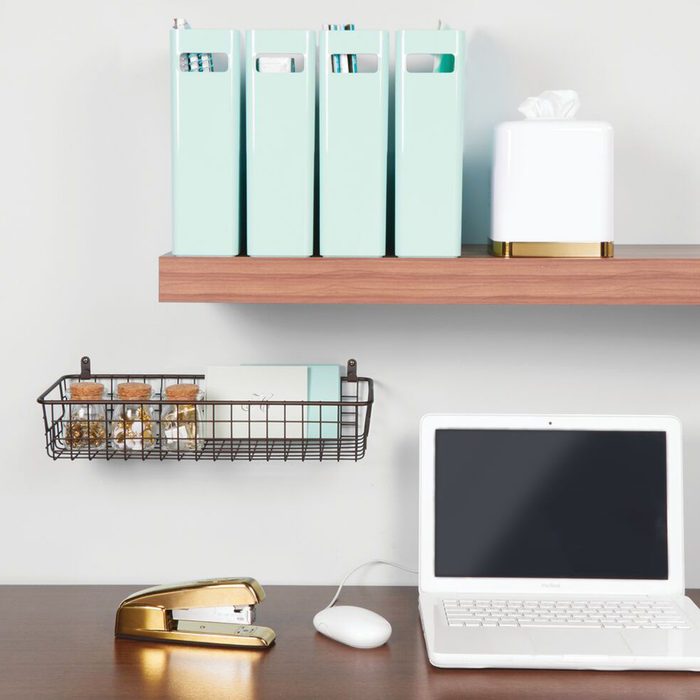 Four mint-colored storage boxes sit on a wooden shelf above a wire basket and a white laptop, with a stapler and mouse on the desk.