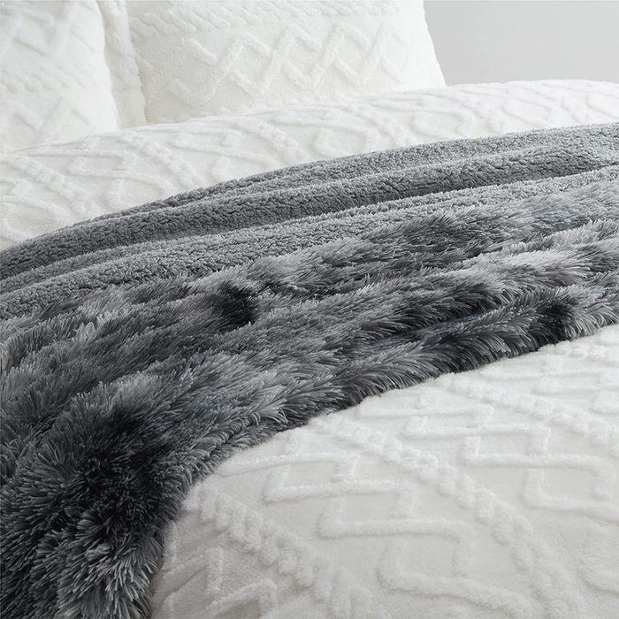 A soft, gray, fuzzy throw blanket rests across a textured white blanket on a bed, creating a cozy appearance in a serene bedroom setting.