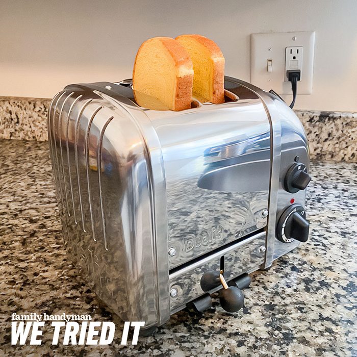 A stainless steel toaster sits on a granite countertop, toasting two slices of bread. A power outlet is visible behind it.
