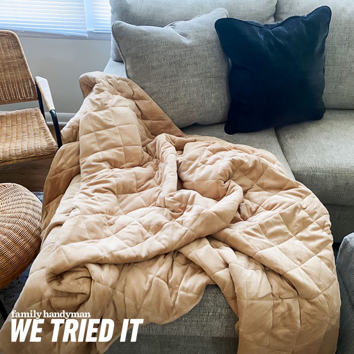 A cozy, beige blanket lies rumpled on a gray sofa, alongside a dark cushion and a woven chair, bathed in natural light from nearby windows.