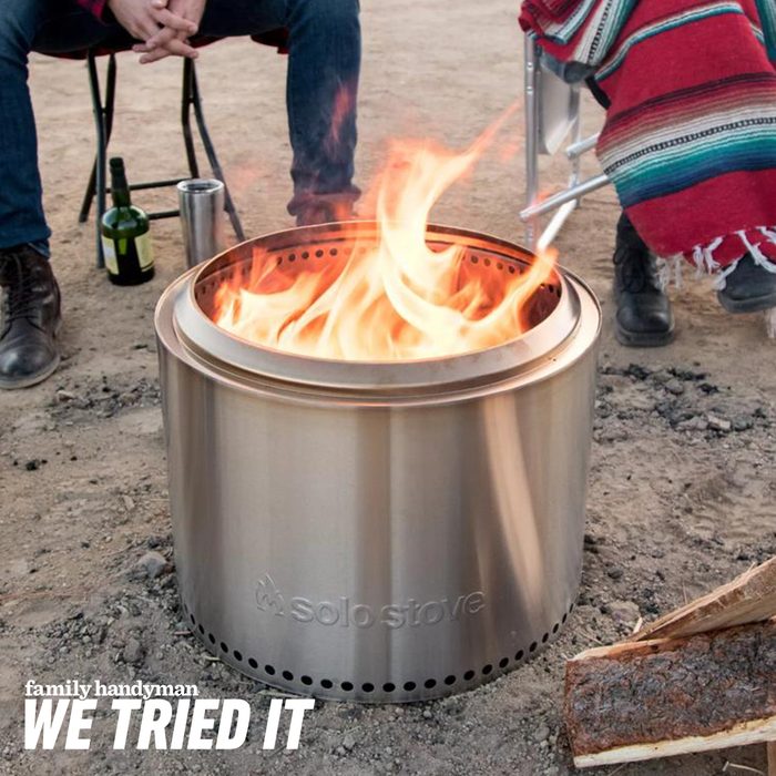 A stainless steel fire pit is glowing with flames, surrounded by seated individuals on dirt, with a bottle and tumbler nearby.