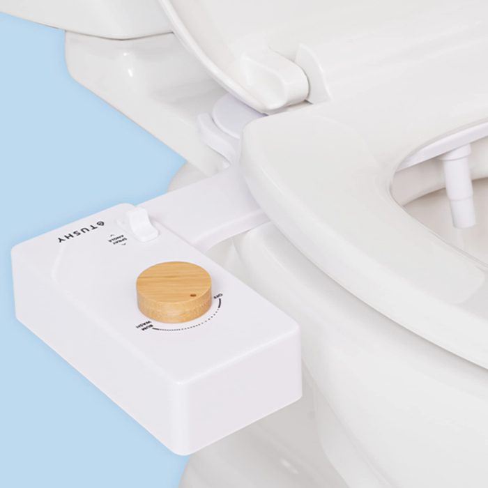 A toilet bidet attachment is shown, featuring a wooden dial for adjusting settings, positioned beside the toilet seat against a light blue background.