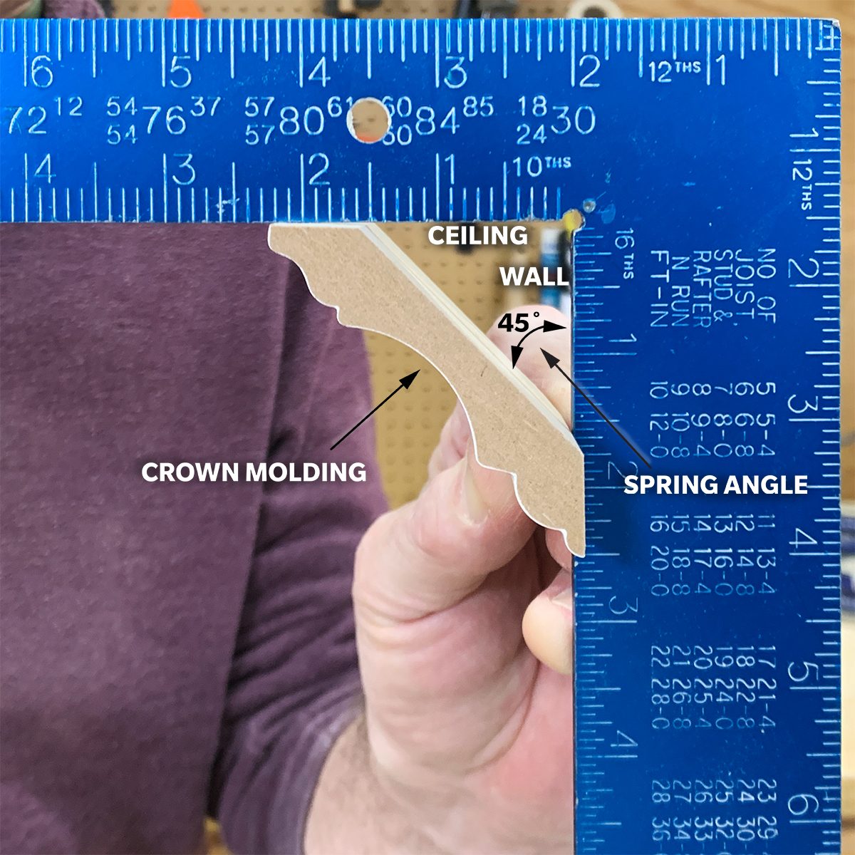 A person holds a piece of crown molding against a blue ruler marked with angles, illustrating a technique in a workshop setting.