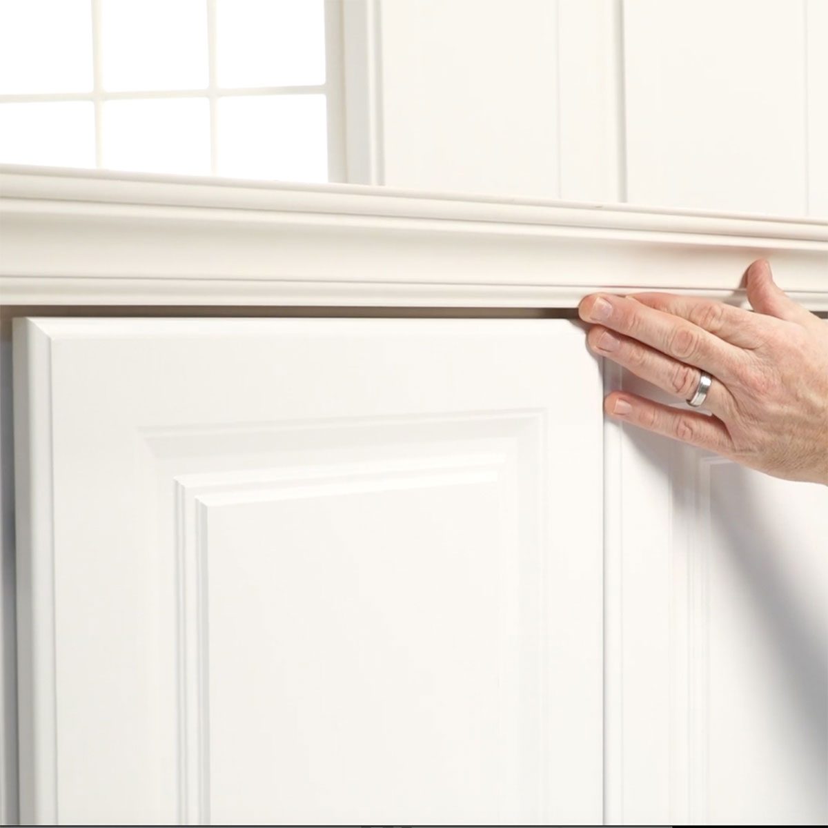 A hand touches the edge of a white cabinet door, aligning it with a decorative trim in a bright, well-lit room.