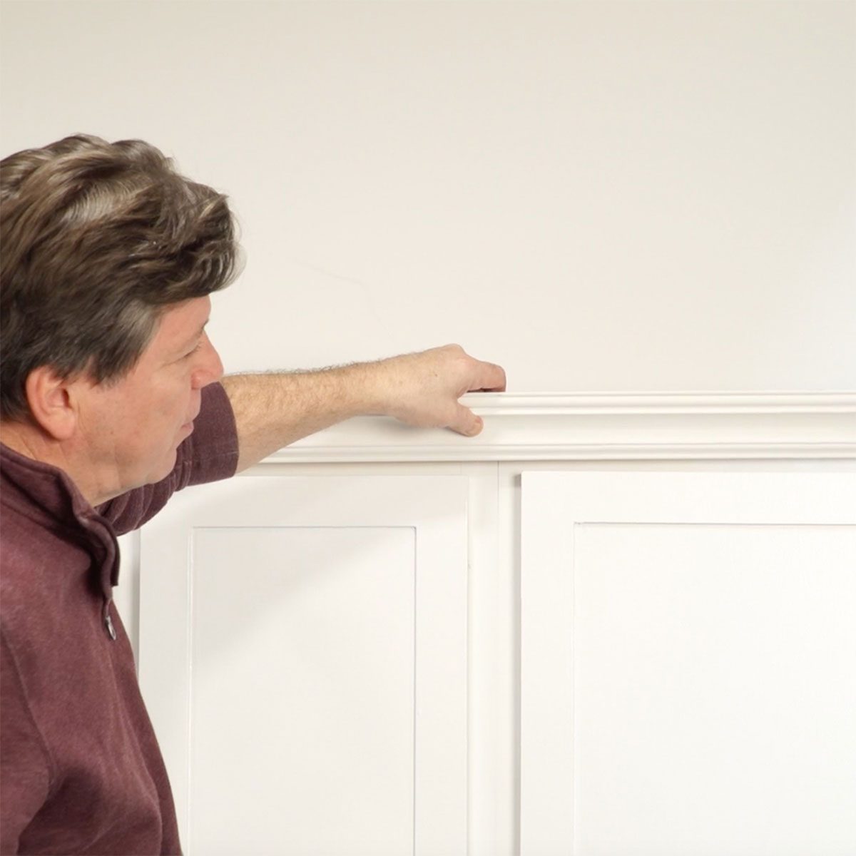 A man points to a white trim above a smooth wall, engaged in measuring or discussing its placement in a bright, uncluttered space.