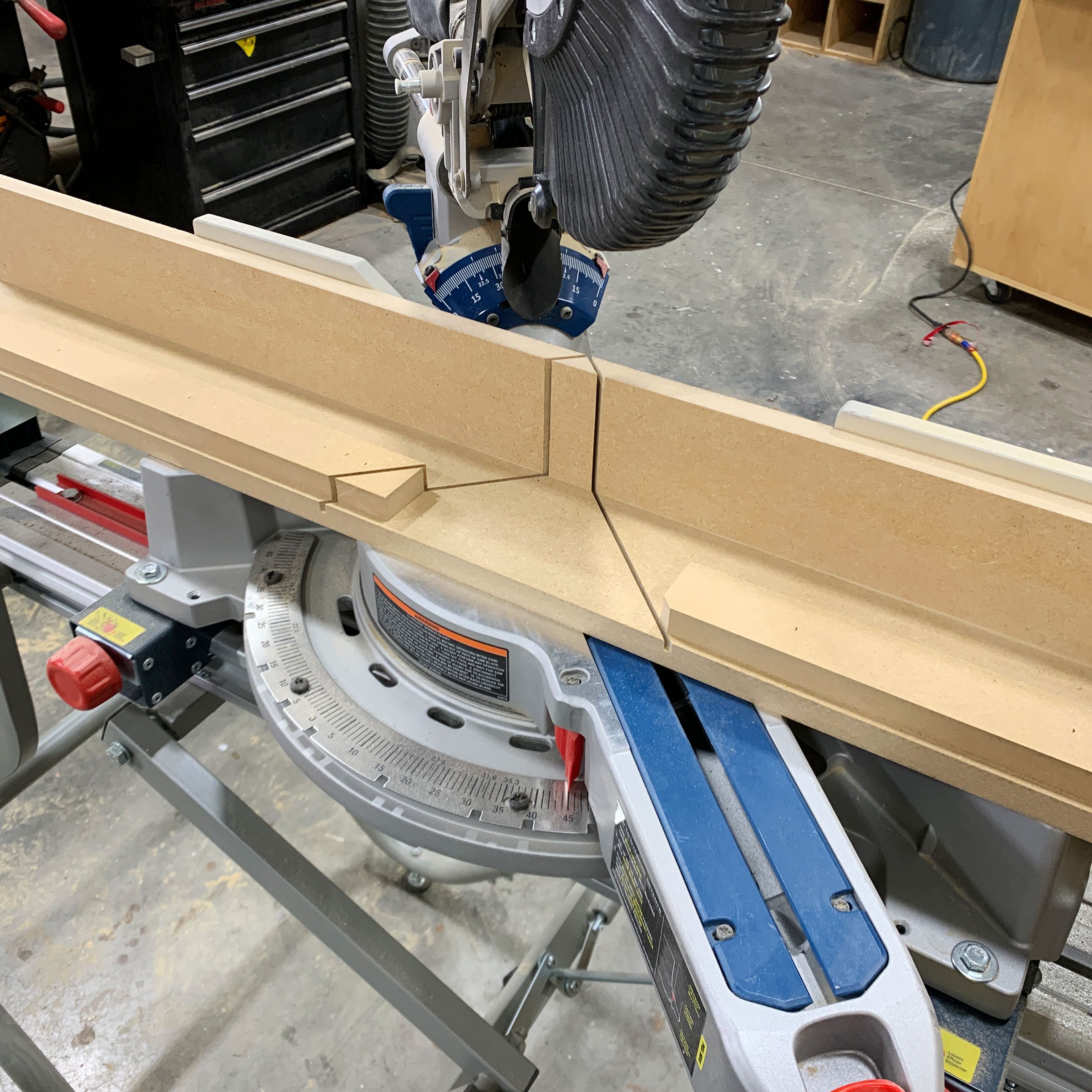 A miter saw sits on a workbench, cutting through a piece of MDF, surrounded by tools and shop equipment in a workshop environment.