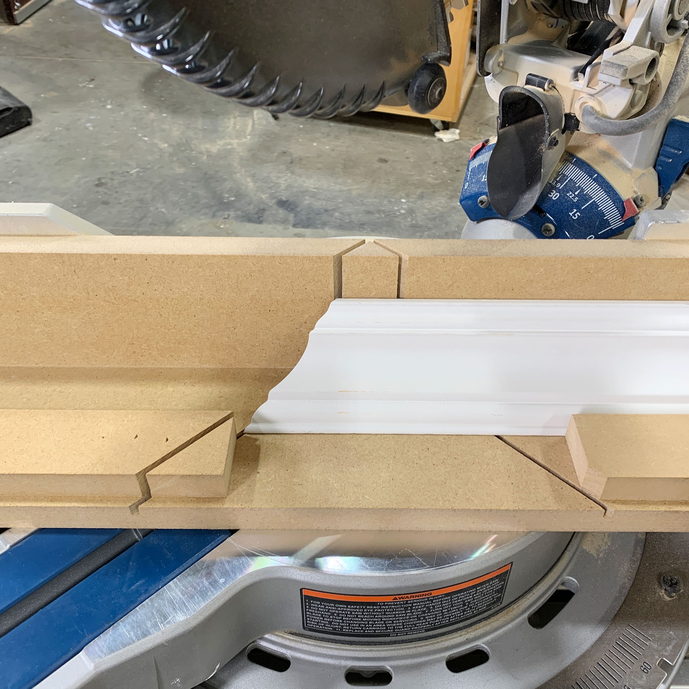 A saw blade hovers above a workpiece, positioned on a miter saw with wooden guide blocks, indicating preparation for cutting in a workshop setting.