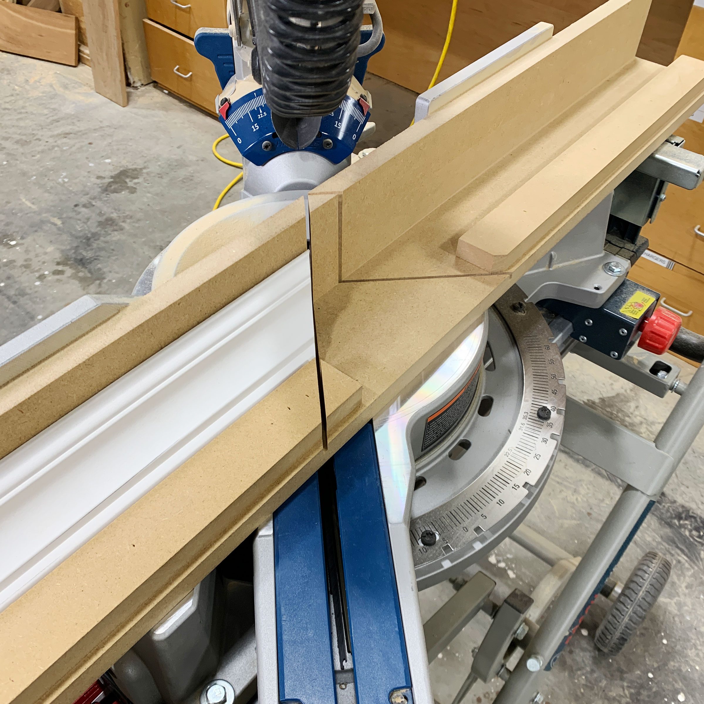 A miter saw is cutting a piece of molding, angled for precise fitting. The workspace features tools and storage in the background, suggesting a carpentry shop.