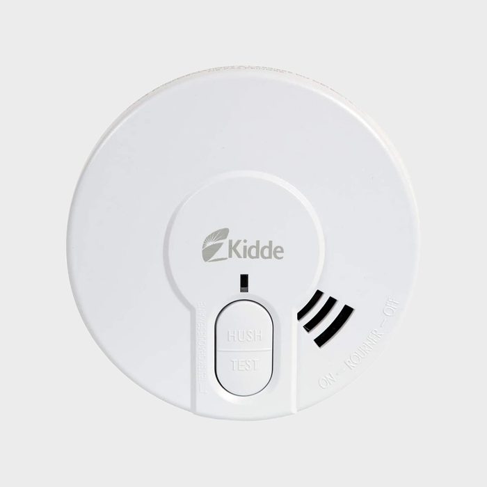 A white smoke detector is mounted, featuring buttons labeled “HUSH” and “TEST,” with a logo and indicator lights, against a plain background.