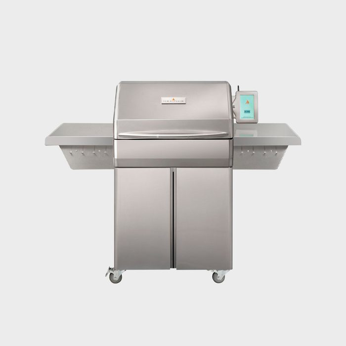 A stainless steel grill stands vertical, featuring a lid and side tables. It's designed for cooking outdoors, evident by its robust, wheeled base for mobility.