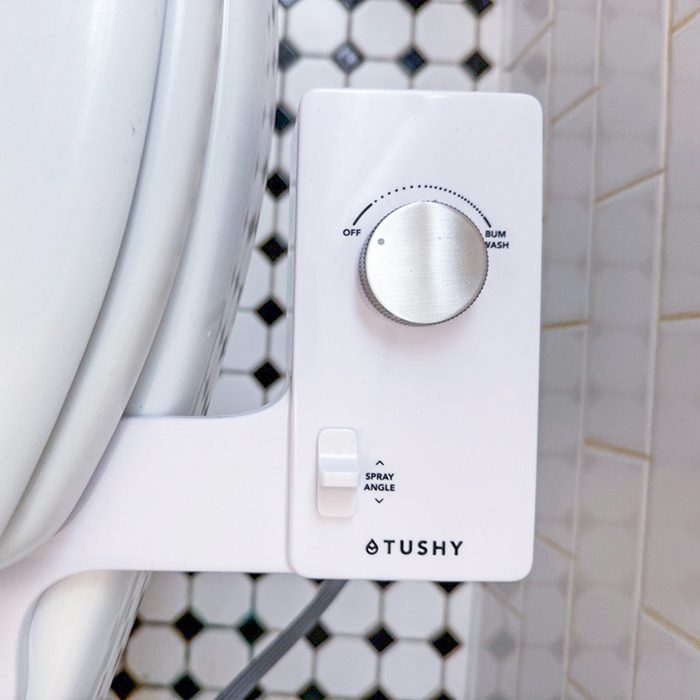 A white bidet attachment with knobs for spray angle and wash settings is mounted beside a toilet, set against a tiled wall pattern.