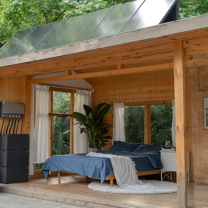 A compact wooden bedroom features a bed with blue linens, standing near windows and plants, surrounded by greenery and natural light, with solar panels overhead.