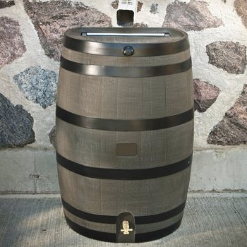 A wooden rain barrel sits against a textured stone wall, equipped with a spout at the bottom for water access.