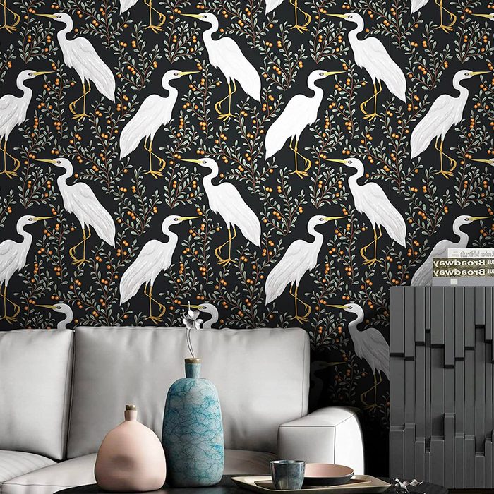 A patterned wallpaper features white herons among colorful foliage. A modern couch and decorative vases sit in front, enhancing a stylish indoor space.