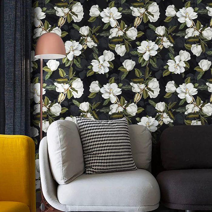 A light gray sofa with a black-and-white patterned pillow sits against a floral wallpaper background, with a yellow chair and a lamp nearby.