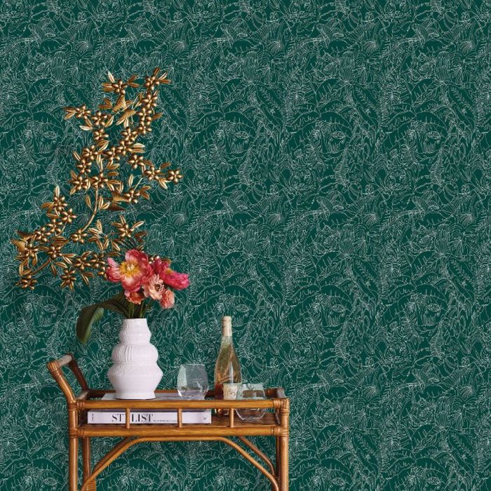 A decorative table holds a white vase with colorful flowers, alongside glasses and a bottle. Behind, a green patterned wallpaper adds texture to the scene.