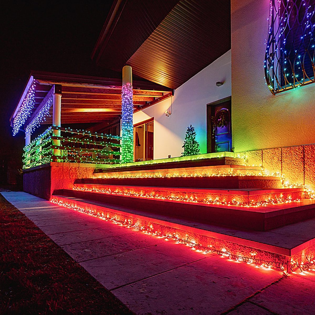 Colorful holiday lights illuminate a staircase and porch, creating a festive atmosphere in a residential setting during nighttime. A small Christmas tree adds to the decor.