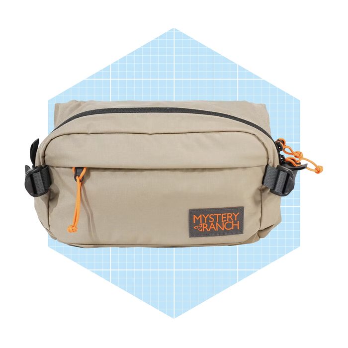 A beige bag with an orange zipper pull sits against a light blue, grid-patterned background, showcasing its streamlined design and brand label.