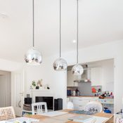 Three metallic pendant lights hang above a wooden dining table in a bright, modern living space featuring a kitchen and entertainment area.