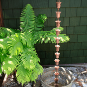 A copper rain chain hangs vertically, directing water into a round metal basin. Lush green ferns frame the scene against a textured green wall.