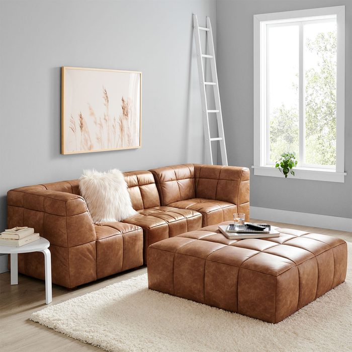 A brown leather sectional sofa sits on a plush rug, accompanied by a square ottoman. A wooden ladder leans against a gray wall near a bright window.