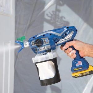 A blue airless handheld paint sprayer sprays liquid, held by a hand, against a light surface, surrounded by a protective covering.