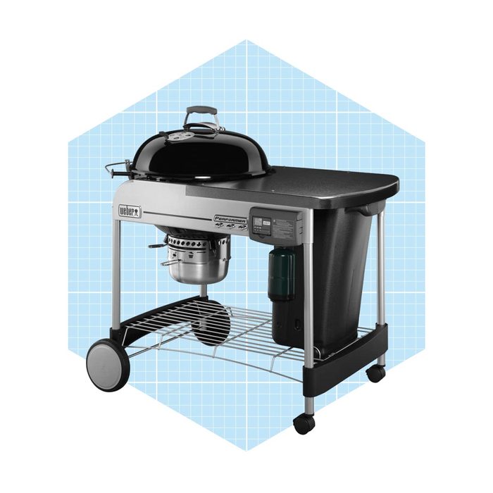 A black kettle grill sits on a cart with wheels, featuring a side table, storage space, and a propane tank holder against a light blue grid background.
