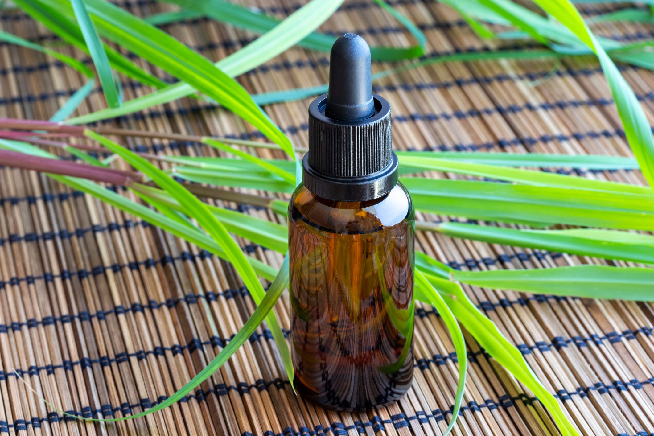A glass dropper bottle sits on a woven mat, surrounded by green leaves, suggesting a natural or herbal context.