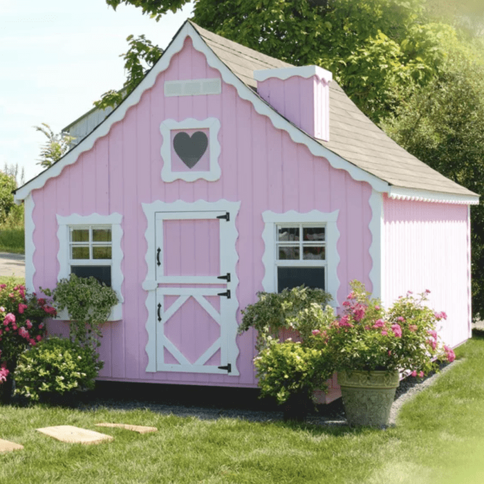 A pink cottage with white trim sits surrounded by green grass and colorful flowers, featuring a heart-shaped window and a decorative door.