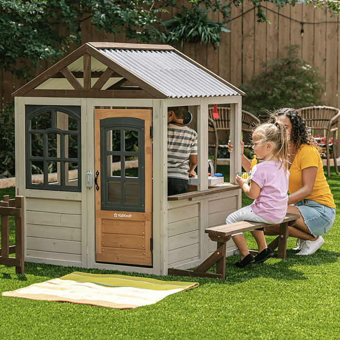 A playhouse stands in a green backyard. Two children interact at a play kitchen, while an adult sits nearby, watching and engaging with them.