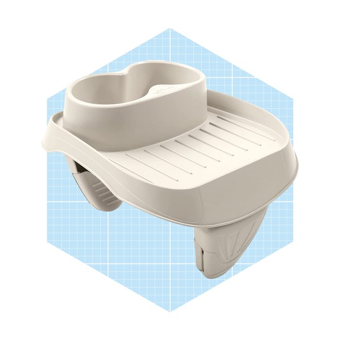 A beige baby bath seat is displayed against a light blue grid background. It features a raised edge and grips for secure placement.