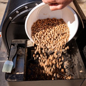 A hand pours wood pellets from a white bowl into a pellet grill’s hopper, surrounded by a black grill in an outdoor setting.