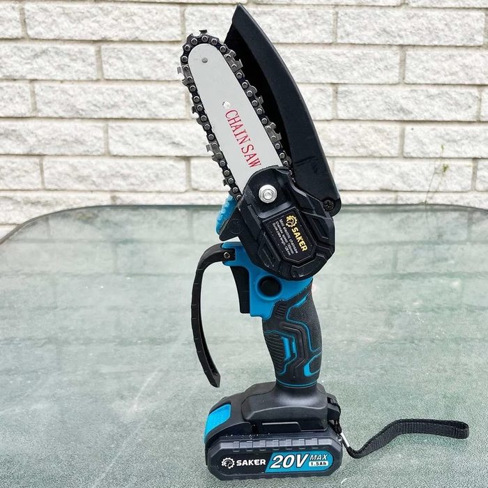 A handheld chainsaw rests on a glass table, featuring a chain and a battery pack, set against a textured brick wall background.