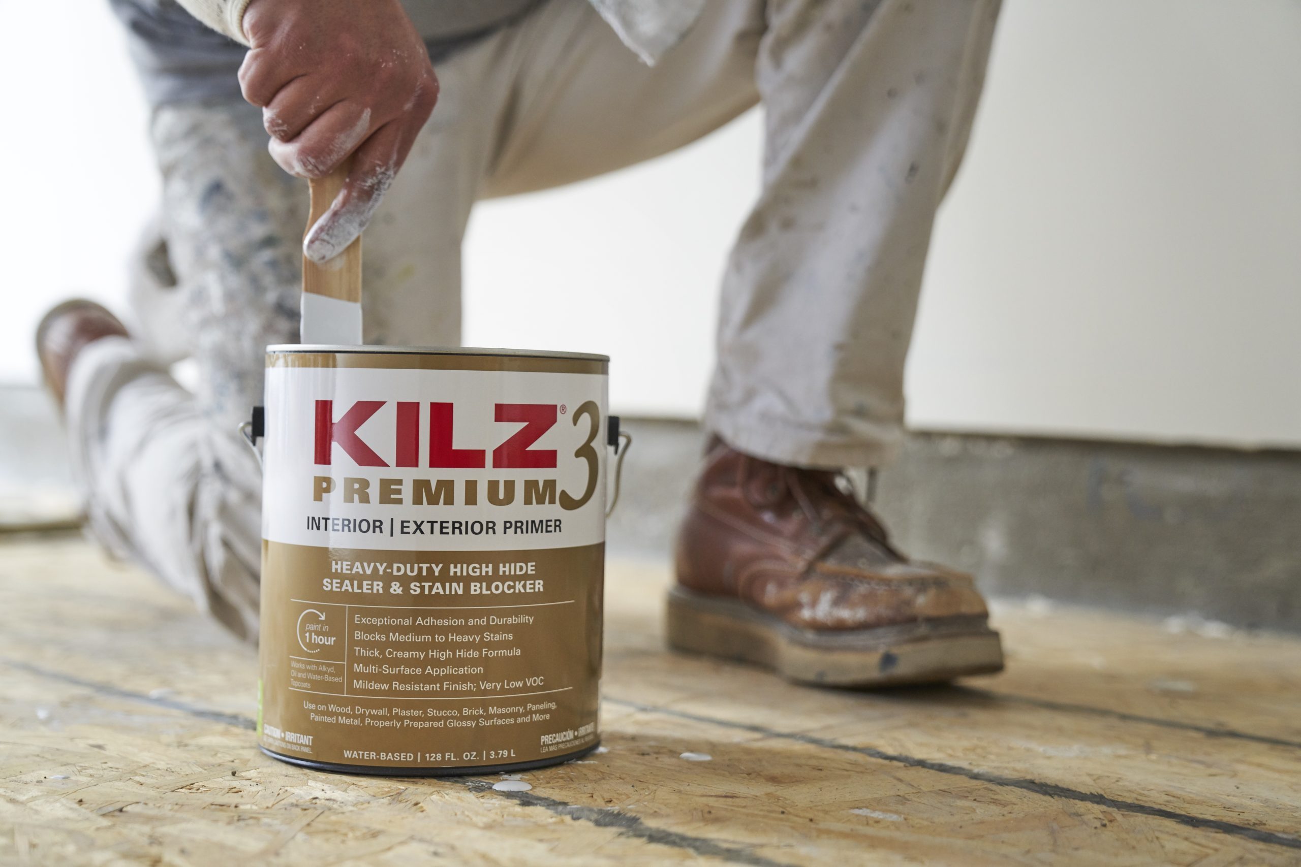 A painter kneels, stirring a can of primer with a brush on a wooden floor, surrounded by an unfinished room. Brown work boots are visible.